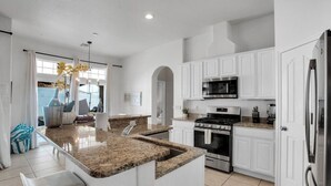 A modern kitchen with white cabinetry, a granite countertop island, stainless steel appliances, and a large window. - The Desert Gem Pool Spa Gated Private Home (Desert Hot Springs)