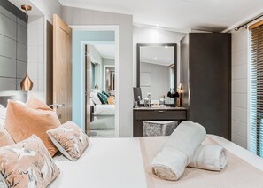 A modern hotel room with a large bed, a bedside table, a mirror, and a view into the adjacent room.