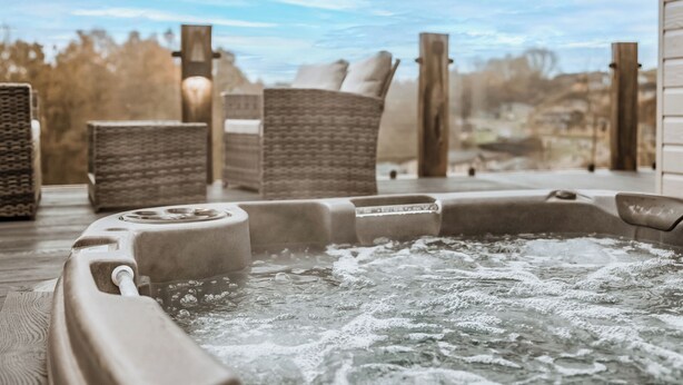Een hottub met een houten terras, rieten stoelen en uitzicht op bomen en een gebouw in de verte.