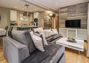 Ein modernes Wohnzimmer mit einem grauen Ecksofa, einem Flachbildfernseher und einem Essbereich, wobei in der Ferne eine Küche zu sehen ist.