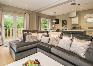 A modern living room with a sectional sofa, a dining area, and a kitchen. - Deluxe lodge with one master double with an en suite shower and one twin. (Cartmel)