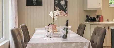 Una mesa de comedor con sillas, una araña, una orquídea en maceta, una botella de champán y un cuadro de un ciervo.
