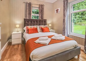 A bedroom with a large bed, two bedside tables with lamps, a window with curtains, and a picture on the wall. - Deluxe lodge with one master double with an en suite shower and one twin. (Cartmel)