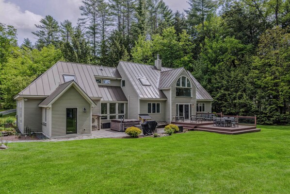 A two-story house with a covered patio, a front deck, and a large yard. - Quechee Meadows Home with Hot Tub & Fire Pit (Quechee)