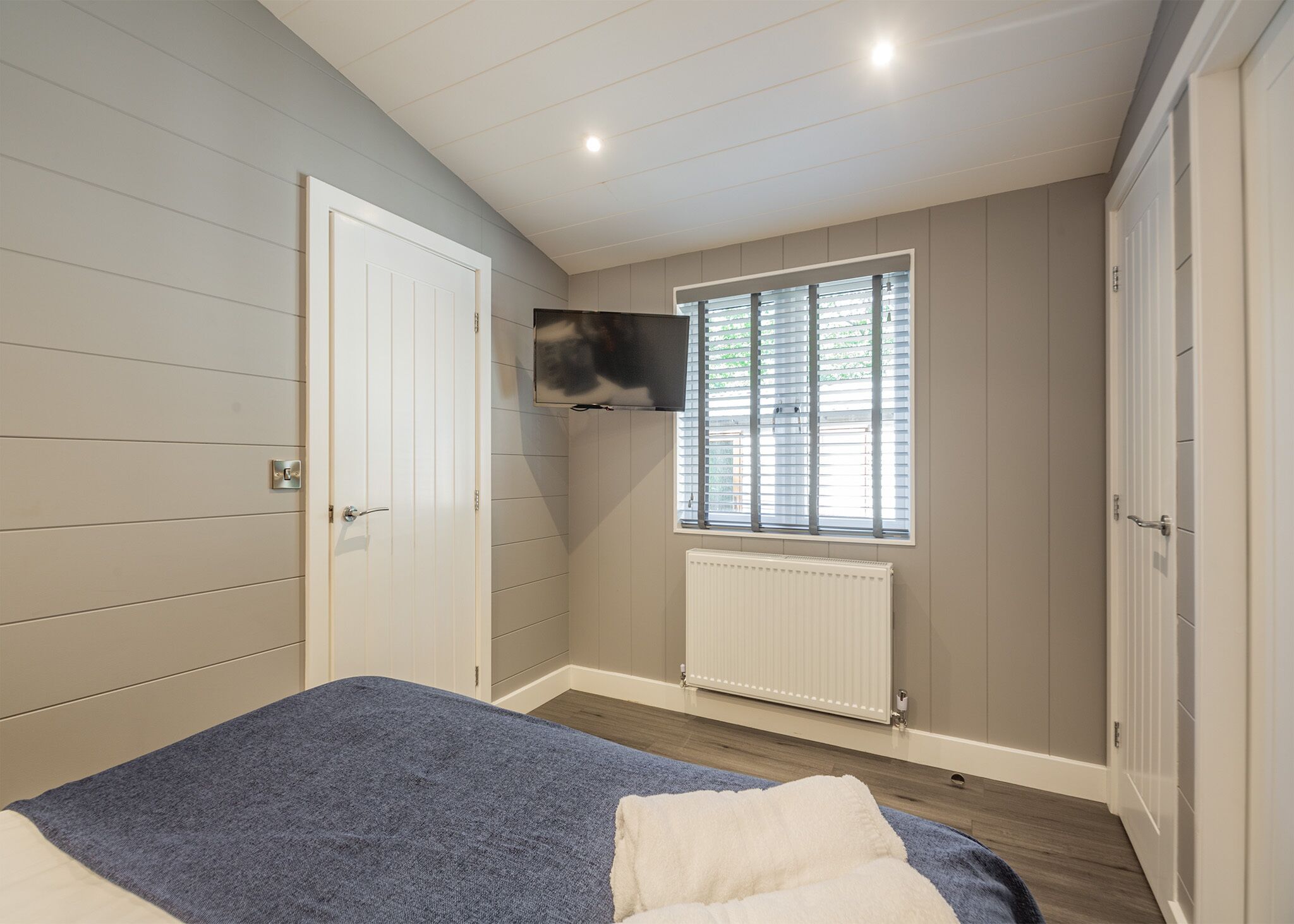 Un dormitorio con cama, televisor montado en la pared, ventana con persianas y radiador.