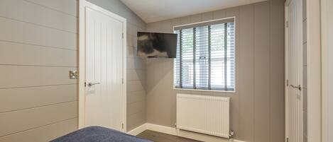 A bedroom with a bed, a TV mounted on the wall, a window with blinds, and a radiator.