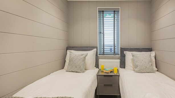 A small, modern bedroom with two single beds, a nightstand, a radiator, and a window with blinds.