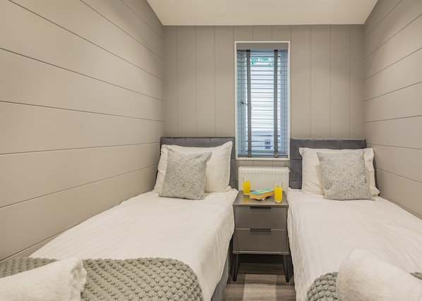 A small, modern bedroom with two single beds, a nightstand, a radiator, and a window with blinds.