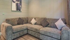 A corner living room with a sectional sofa, patterned cushions, a framed abstract painting, and wooden panel walls.