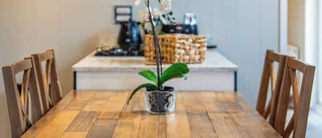 A wooden dining table with chairs, a potted orchid, and a kitchen area in the background.
