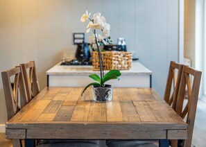 Una mesa de madera con sillas, una orquídea en maceta y una zona de cocina al fondo.