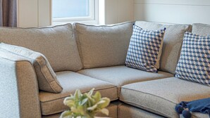A sectional sofa with patterned cushions, a round wooden coffee table with a potted plant, and a window with curtains. - 2 bedroom accommodation in Tewkesbury (Tewkesbury)