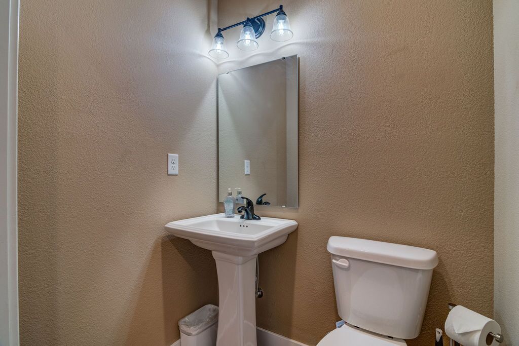A bathroom with a pedestal sink, a toilet, and a mirror with overhead lighting.