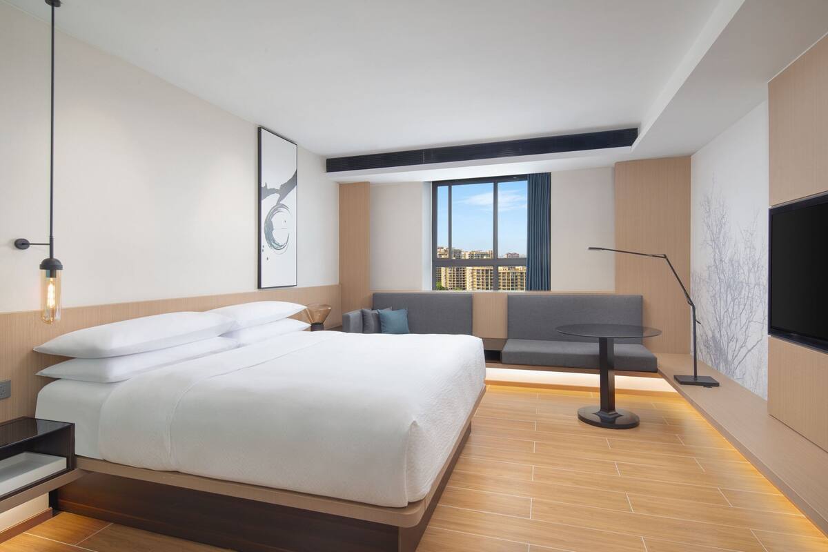 A modern hotel room with a large bed, a seating area with a sofa and a small table, a large window with a city view, and a flat-screen TV mounted on the wall.