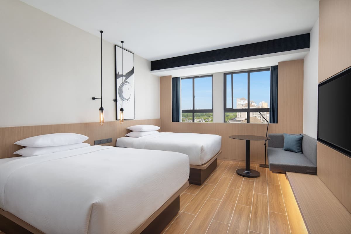 A hotel room with two beds, a sofa, a TV, and a large window offering a city view.