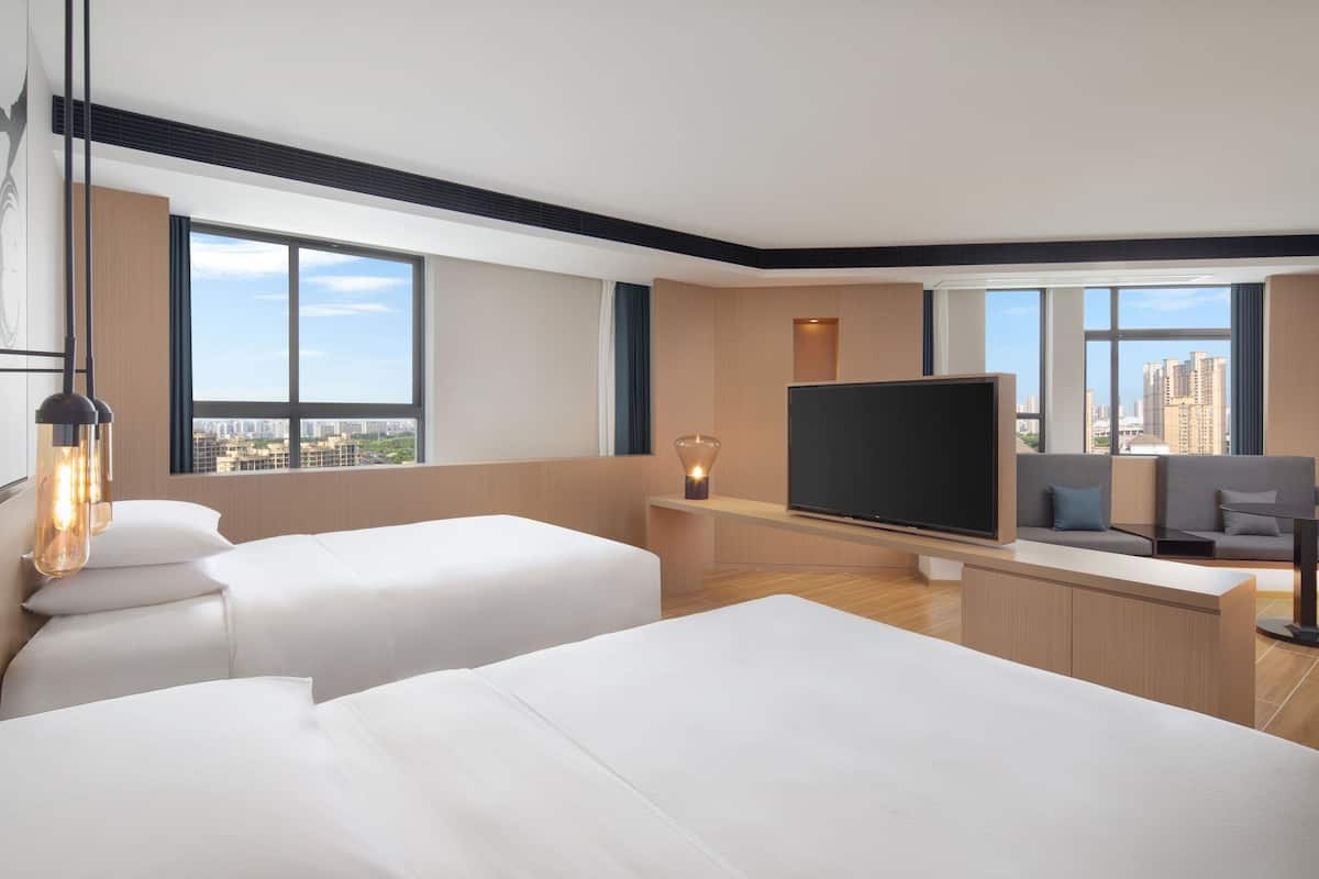 A modern hotel room with two beds, a flat-screen TV, a desk, and a large window offering a city view.