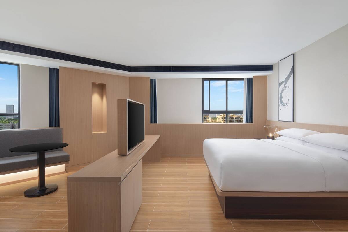 A modern hotel room with a large bed, a TV on a wooden console, a seating area with a bench, and large windows offering a city view.