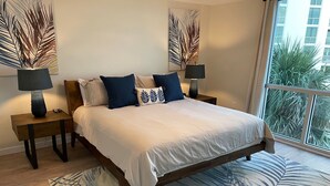 Ein Schlafzimmer mit einem großen Bett, zwei Nachttischen, einer Stehlampe und Blick auf Palmen durch ein Fenster.
