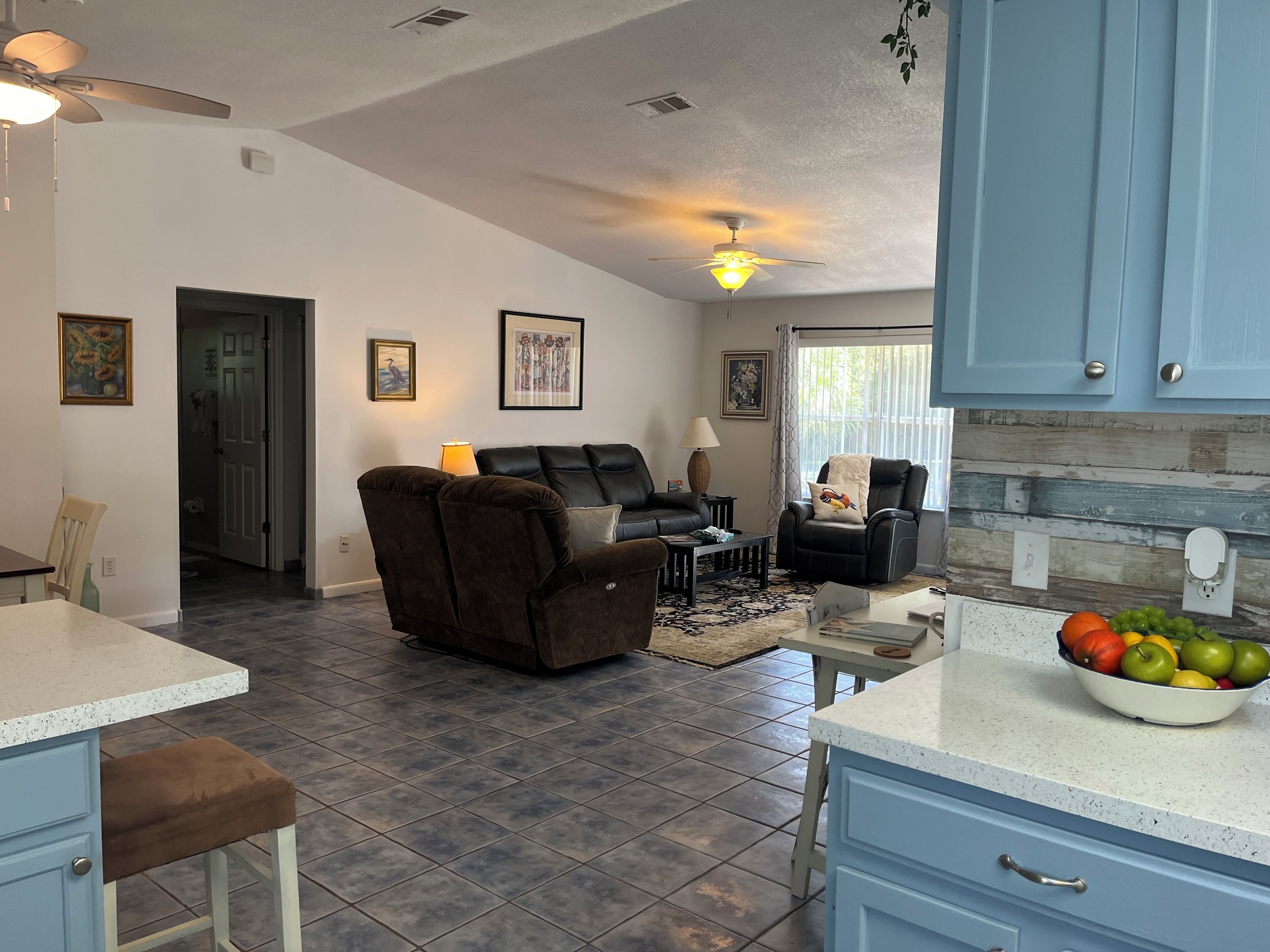 A spacious living area with a dark brown sofa and a black leather chair, a dining area with a white countertop and blue cabinets, and a kitchen with blue cabinets and a fruit bowl.