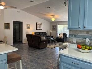 A spacious living area with a dark brown sofa and a black leather chair, a dining area with a white countertop and blue cabinets, and a kitchen with blue cabinets and a fruit bowl. - Family Welcome-Large Comfy Stay Pets Welcome w/Parking Trailers/Bikes/Boats (Edgewater)