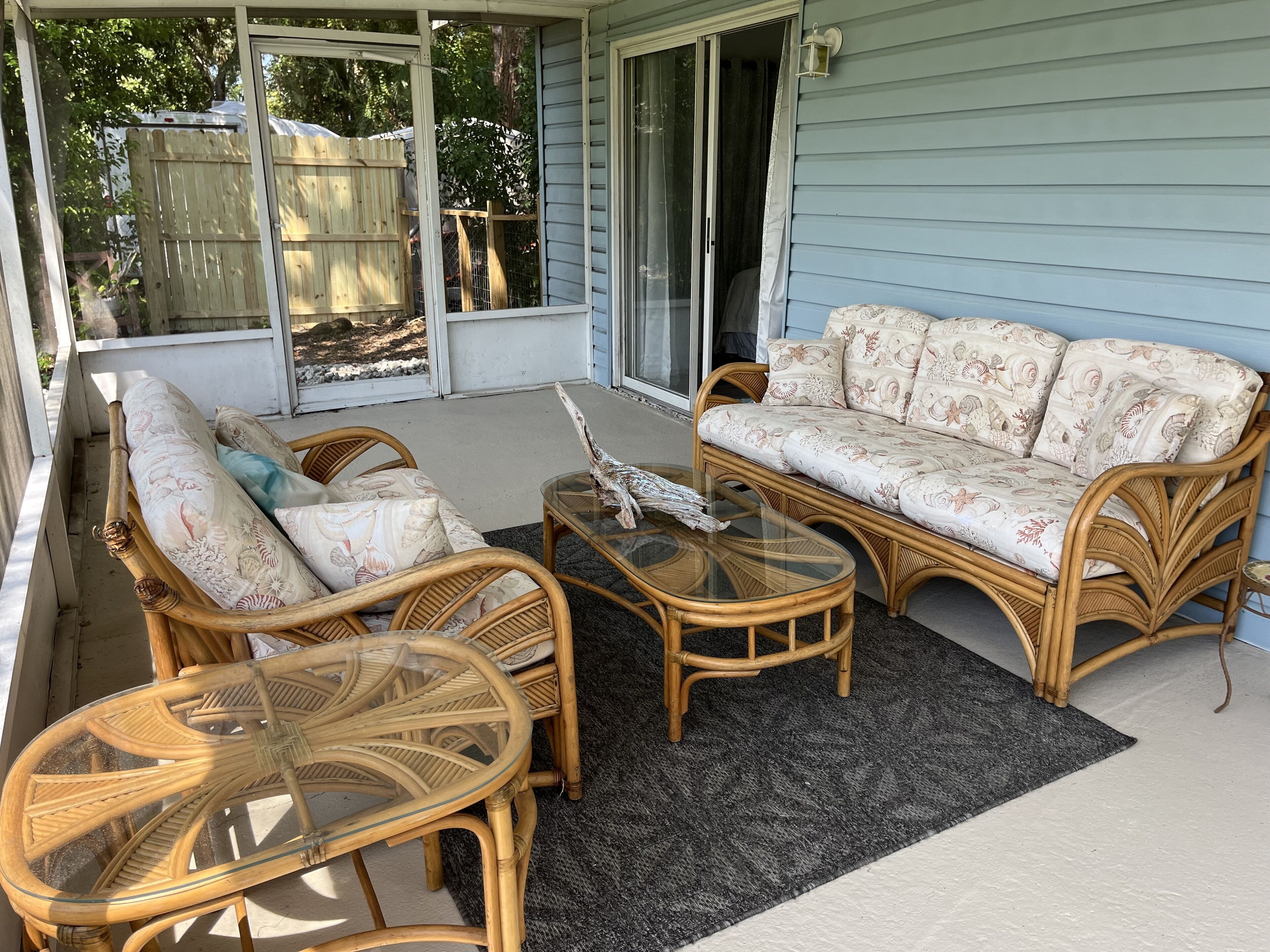 Ein überdachter Patio-Bereich mit Korbmöbeln, darunter eine Couch und zwei Sessel, sowie ein Couchtisch mit Glasplatte.