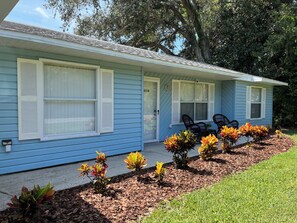 A single-story blue house with white shutters, a front porch with two chairs, and a small garden with colorful plants. - Family Welcome-Large Comfy Stay Pets Welcome w/Parking Trailers/Bikes/Boats (Edgewater)