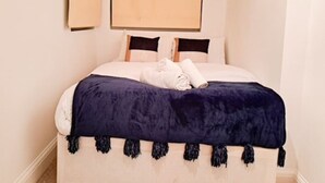 Uma cama bem arrumada com colcha azul escura e franjas, almofadas brancas e toalhas dobradas ao lado.