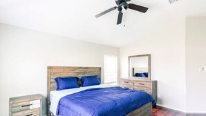 Ein Schlafzimmer mit hölzernem Kopfteil, einem Bett mit blauer Bettwäsche, einem Nachttisch, einer Kommode, einem Deckenventilator und einem Spiegel.