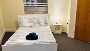 Una cama bien hecha con ropa de cama a rayas y una toalla doblada, una mesita de noche con lámpara y una ventana con persianas.