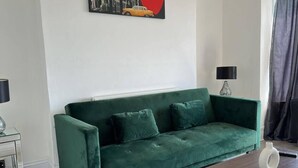A green sofa in a room with a painting of a yellow car on the wall, a lamp, and a vase on a table.