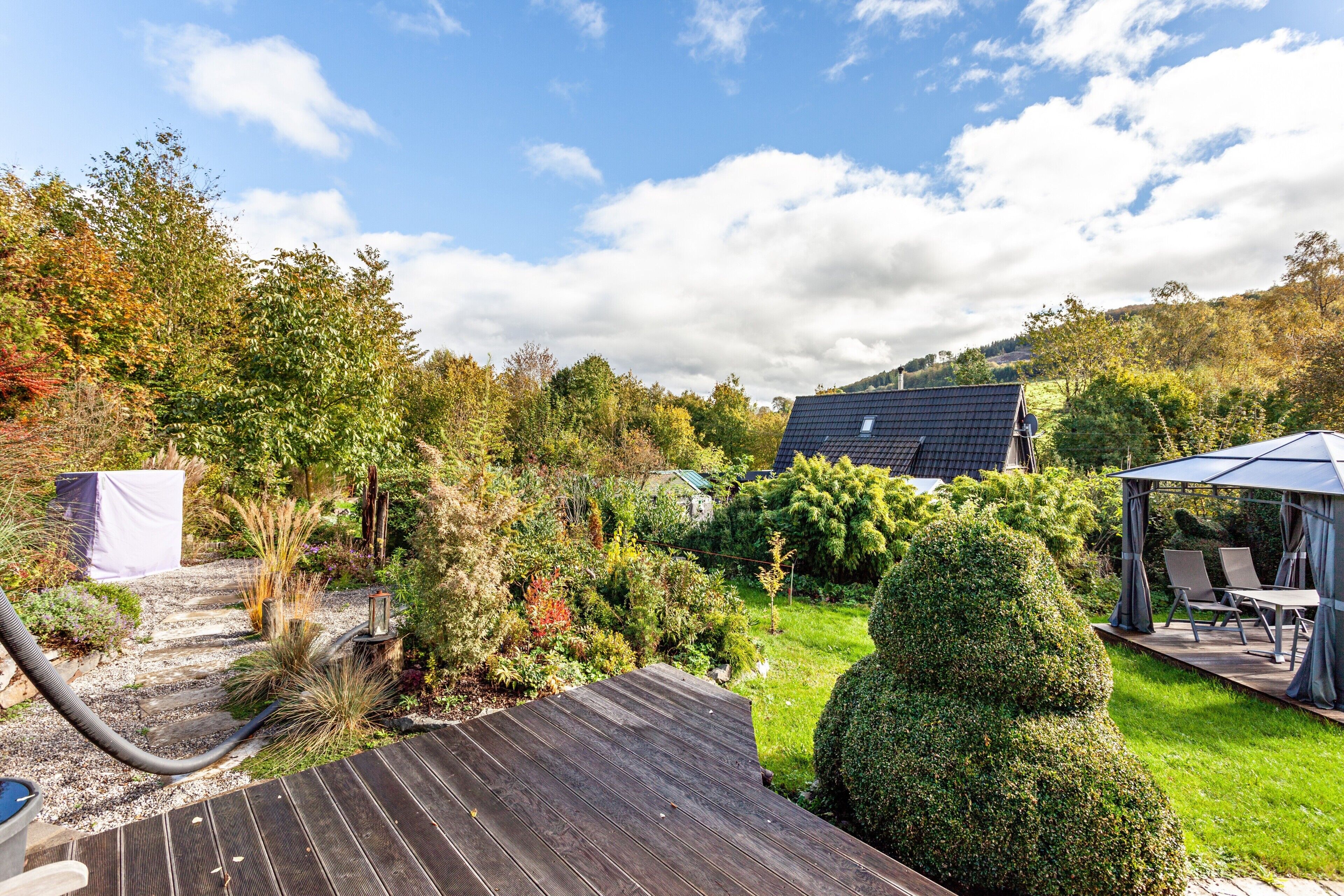 Un jardin avec une terrasse en bois, un belvédère et une maison en arrière-plan.