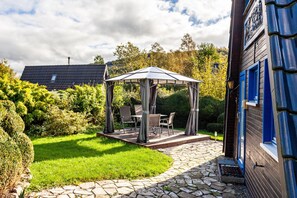 Un belvédère abritant une table et des chaises, entouré d’un chemin de pierre et d’une végétation luxuriante.