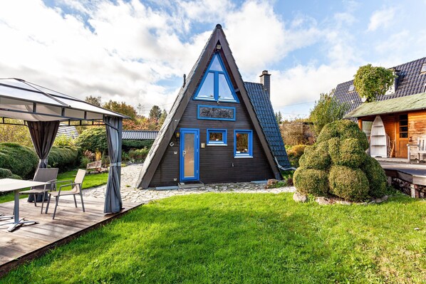 Une maison triangulaire à l’extérieur sombre, agrémentée de touches de bleu, une terrasse avec des meubles d’extérieur et un jardin bien entretenu.