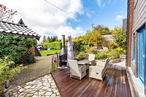 Une terrasse en bois avec des meubles d’extérieur, un chemin en pierre et un espace jardin agrémenté d’une cheminée.