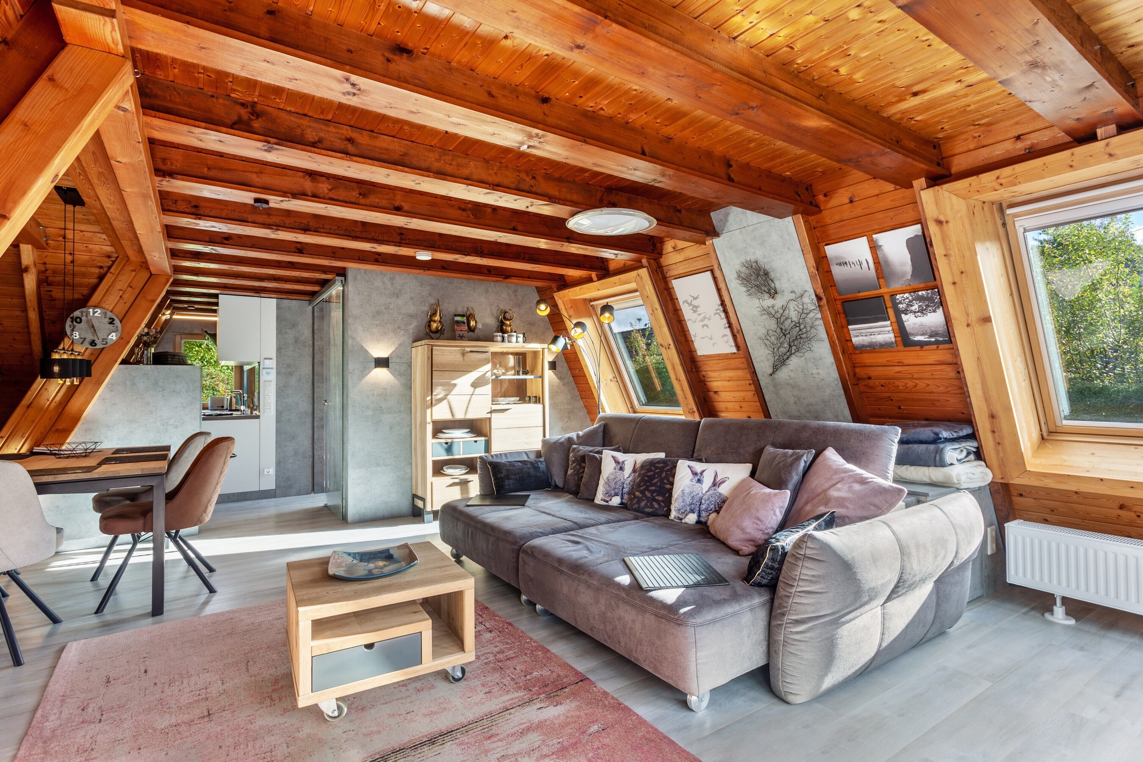 A cozy cabin interior with a wooden ceiling, a sofa, a dining table, and a view through a window.