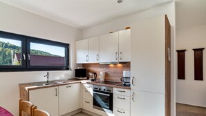A compact kitchen with white cabinets, a built-in oven, a coffee maker, and a window with a view of a forest.