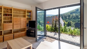 A modern living room with a large sliding glass door to a garden, a wooden bookshelf, and a flat-screen TV.
