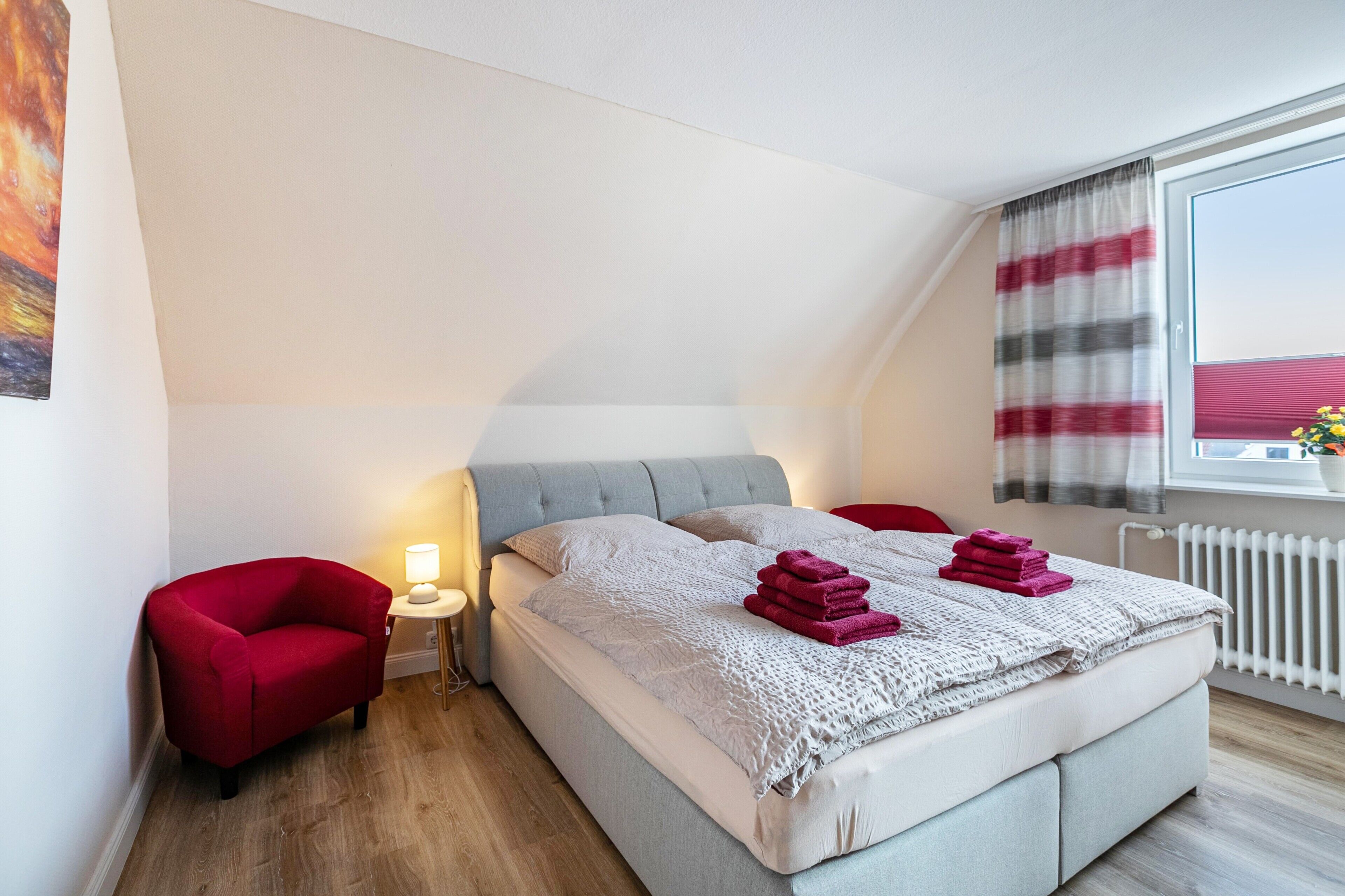 Ein Schlafzimmer mit einem Bett, einem roten Sessel, einem Nachttisch mit Lampe und einem Fenster mit Vorhängen.