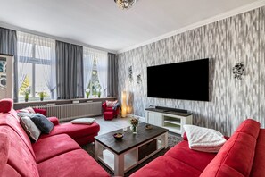 A living room with a red sofa, a coffee table, a flat-screen TV mounted on the wall, and a window with curtains.