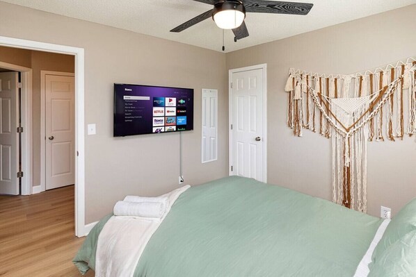 A bedroom with a bed, a ceiling fan, a wall-mounted TV displaying streaming services, and a macrame wall hanging.