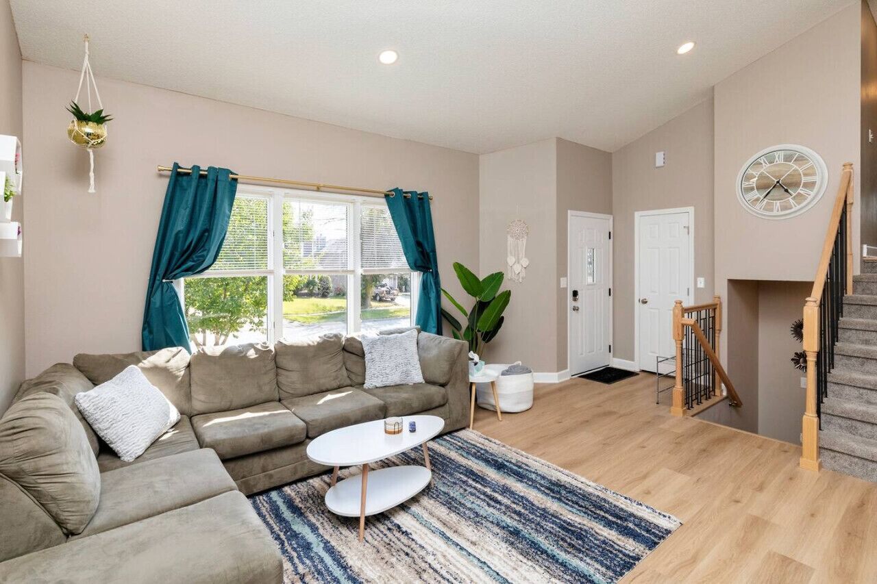 A living room with a sectional sofa, a coffee table, a large window with curtains, a staircase, and a clock on the wall.