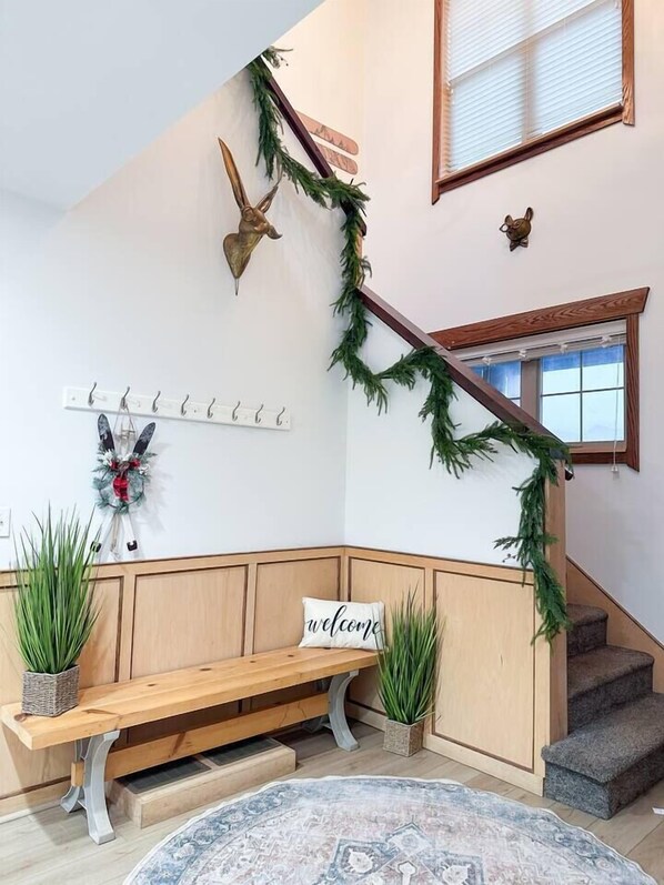 A staircase with a wooden handrail, a bench with a 'welcome' sign, a coat rack with hooks, and a decorative wall with a deer head and greenery. - SKI IN/OUT- Pool table, hot tub, A+ kitchen, 7 bedrooms->4 kings+custom kid rms! (Snowshoe)