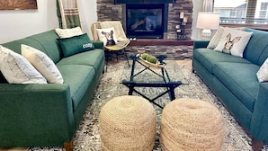 A cozy living room with two green sofas, a fireplace, a flat-screen TV, and a wicker coffee table. - SKI IN/OUT- Pool table, hot tub, A+ kitchen, 7 bedrooms->4 kings+custom kid rms! (Snowshoe)