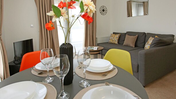 Ein Essbereich mit einem Tisch für vier Personen, einer Blumenvase und einem grauen Sofa im Hintergrund.