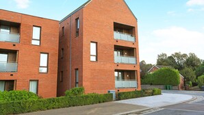 Ein modernes Mehrfamilienhaus aus Backstein mit je zwei Balkonen pro Etage, einer gepflasterten Auffahrt und einem gepflegten Heckenstreifen vorne.