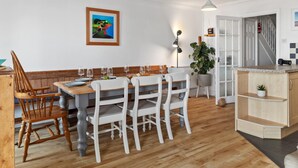 A dining area with a wooden table and white chairs, a framed painting on the wall, and a potted plant near the door. - Penrose - Beach House With Direct Beach Access (Saundersfoot)
