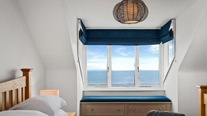 A bedroom with a large window, a wooden headboard, a bedside table, and a bed with white bedding. - Penrose - Beach House With Direct Beach Access (Saundersfoot)