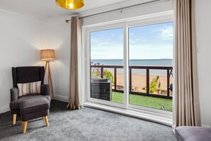 A room with a large window offering a view of the beach, a chair, a floor lamp, and a balcony with a railing.