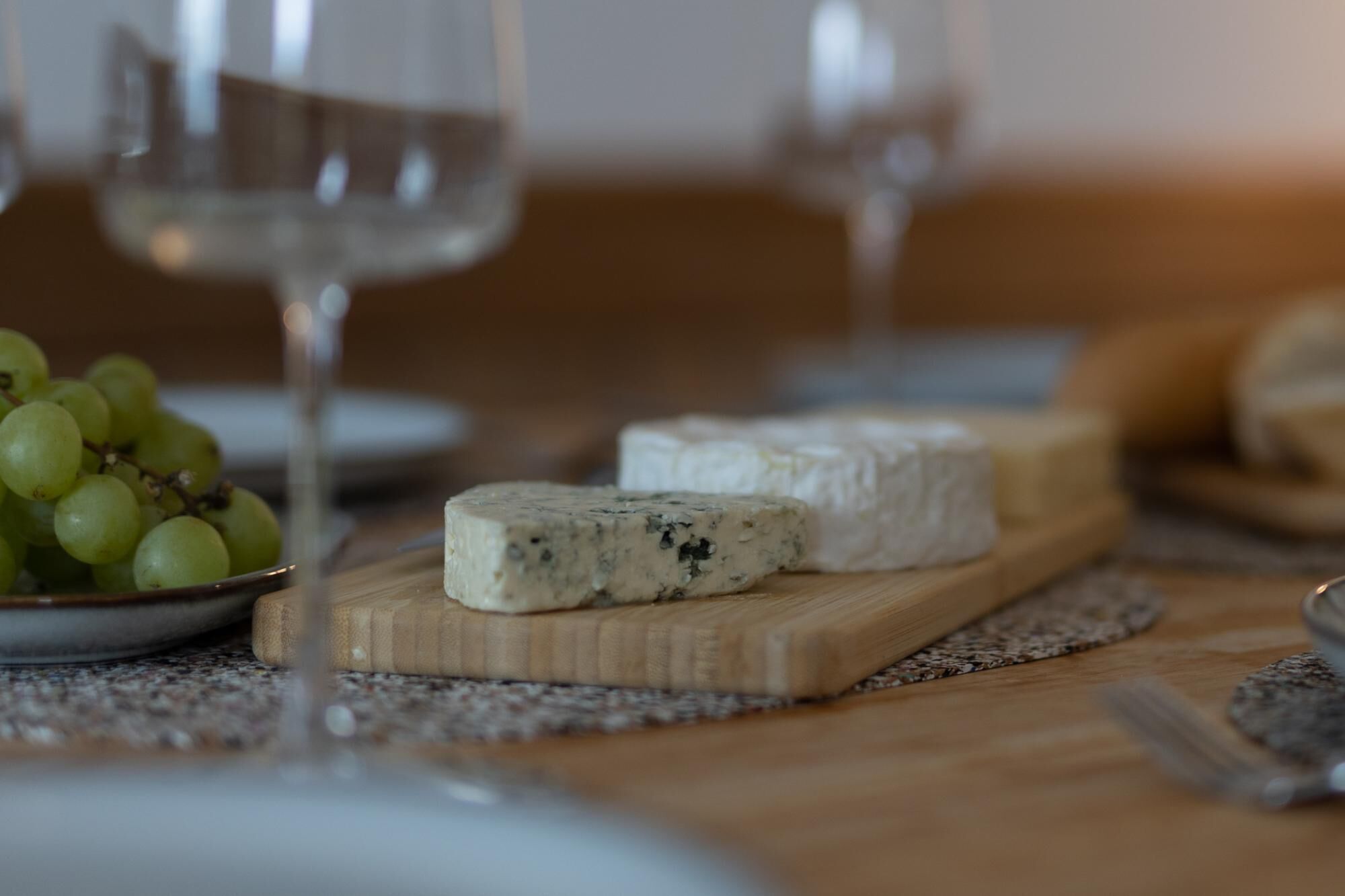 A wooden table with a cutting board holding a piece of blue cheese and a bowl of green grapes.