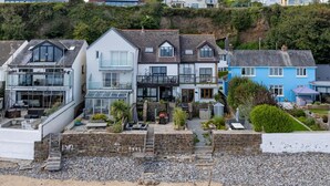 A coastal residential area with houses featuring balconies, a sandy beach in the foreground, and a rocky cliff in the background. - Penrose - Beach House With Direct Beach Access (Saundersfoot)